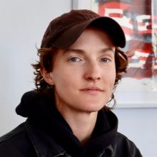 Headshot of the playwright, a white person in a hat with short dark brown hair.